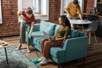 Woman using a laptop on a teal couch while two people clean in the background.